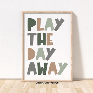 May include: A framed print with the text "PLAY THE DAY AWAY" in green, grey, and beige colors. The text is in a playful, hand-drawn style. The frame is a light wood color.