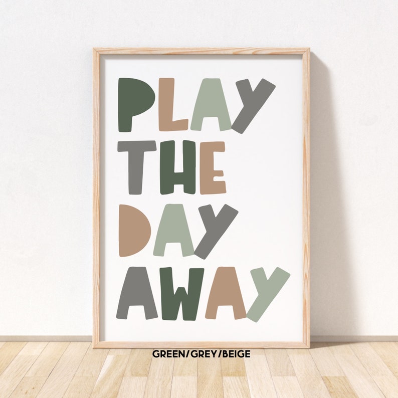 May include: A framed print with the text "PLAY THE DAY AWAY" in green, grey, and beige colors. The text is in a playful, hand-drawn style. The frame is a light wood color.
