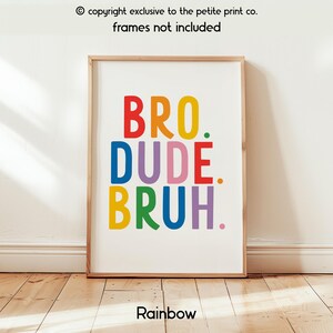 May include: A framed print with the words "BRO. DUDE. BRUH." in rainbow colors. The print is in a wooden frame and is displayed against a white wall. The word "Rainbow" is at the bottom of the print.