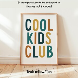 May include: A rectangular wall art print featuring the text COOL KIDS CLUB in a bold, sans-serif font. The letters are colored in alternating shades of teal, mustard yellow, and tan against a white background. The print is housed in a light wood frame.