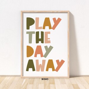 May include: A framed print with the text "PLAY THE DAY AWAY" in colorful, stylized letters. The letters are in shades of green, orange, pink, and brown. The print has a white background and a wooden frame. The word "BOHO" is printed in black at the bottom of the print.