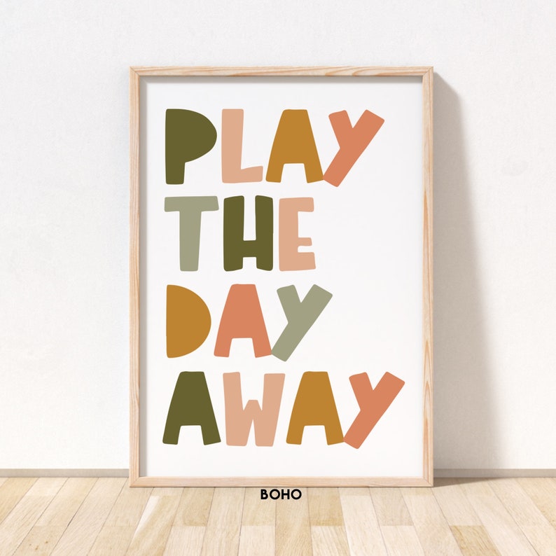 May include: A framed print with the text "PLAY THE DAY AWAY" in colorful, stylized letters. The letters are in shades of green, orange, pink, and brown. The print has a white background and a wooden frame. The word "BOHO" is printed in black at the bottom of the print.