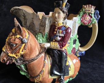 Puede incluir: Una tetera de cerámica decorativa que representa a una mujer con uniforme militar a caballo. La tetera es marrón, con un asa floral y una corona. El caballo tiene arneses dorados y la escena se desarrolla frente a un edificio.