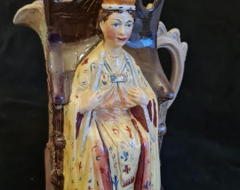 Puede incluir: Una jarra de cerámica con una figura detallada de una reina que lleva una corona y una túnica. La jarra está decorada con pintura marrón, amarilla, roja y azul.