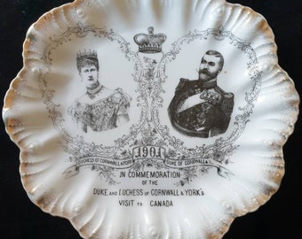 Puede incluir: Un plato de cerámica blanca con un borde dorado presenta una imagen en blanco y negro del duque y la duquesa de Cornualles y York. El plato conmemora su visita a Canadá en 1901. El texto dice "1901, Duquesa de Cornualles y York, Duque de Cornualles y York, En conmemoración de la visita del duque y la duquesa de Cornualles y York a Canadá."