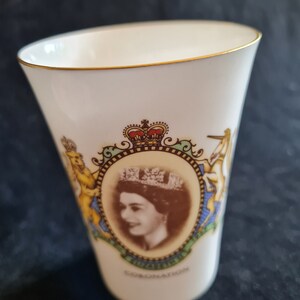 Op de afbeelding: Witte keramische mok met een gouden rand. De mok heeft een kleurrijk beeld van koningin Elizabeth II met een kroon en een wapenschild. De tekst "CORONATION" staat gedrukt op de bodem van de mok.