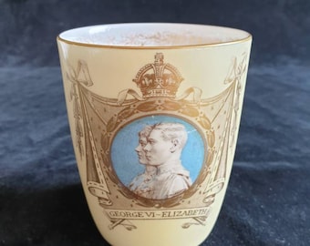 Puede incluir: Una taza de cerámica de color crema con un borde dorado y un diseño dorado que presenta un retrato del rey Jorge VI y la reina Isabel, una corona y el texto "GEORGE VI - ELIZABETH".