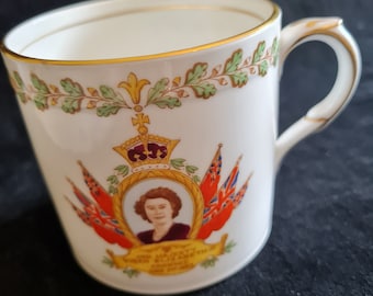 Puede incluir: Taza de cerámica blanca con borde y asa dorados, con un retrato, una corona y banderas. La taza tiene una banda decorativa de hojas verdes y doradas. El texto dice "HER MAJESTY QUEEN ELIZABETH II CROWNED JUNE 2ND 1953."