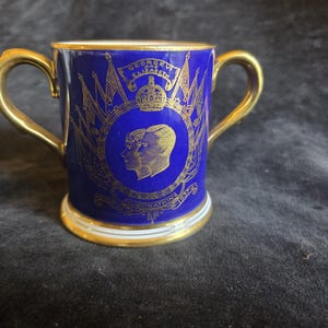 Op de afbeelding: Een blauw en goud herdenkingsmok met twee gouden oren. De mok heeft portretten en tekst, waaronder "George VI Elizabeth Coronation" en een kroon. De rand is goudkleurig.
