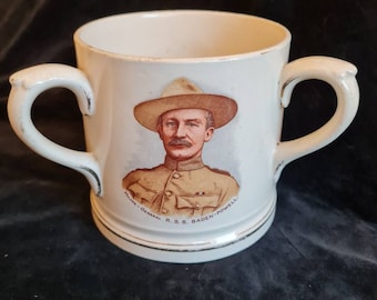 May include: White ceramic mug with two handles, featuring a portrait of Robert Baden-Powell, founder of the Boy Scouts, in uniform and wearing a hat. The text "Major-General R.S.S. Baden-Powell" is printed around the image.