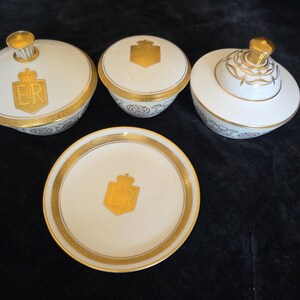 Op de afbeelding: Een set van vier witte porseleinen stukken met gouden randen en accenten. De set bevat twee kommen met deksel, een kleine kom en een bord. Elk stuk heeft een gouden embleem met de letters "ER" en een kroon.