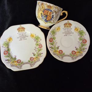 Op de afbeelding: Een vintage theekop en schotel set met een decoratief bloemmotief en gouden rand. De set bevat een theekop, een ronde schotel en een vierkante bord, allemaal met tekst en een koninklijk wapen. De theekop heeft een gouden oor.