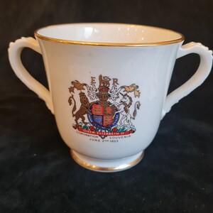 Op de afbeelding: Witte keramische beker met twee oren, gouden rand en een kleurrijk embleem. Het embleem toont een wapenschild en de tekst "Coronation Queen Elizabeth II June 2nd 1953 Souvenir".