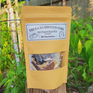 May include: A tan paper packet labeled "THEBACKYARDSEEDSTORE Heirloom Seed Kit Vegetables - 36 Varieties." The packet has a clear window showing various seeds. The background features green plants and a wooden post.