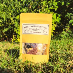 May include: A yellow paper bag with a clear window, filled with seeds. The bag has a white label that reads "THEBACKYARDSEEDSTORE Heirloom Seed Kit Vegetables . 36 Varieties ." The bag is sitting on green grass.