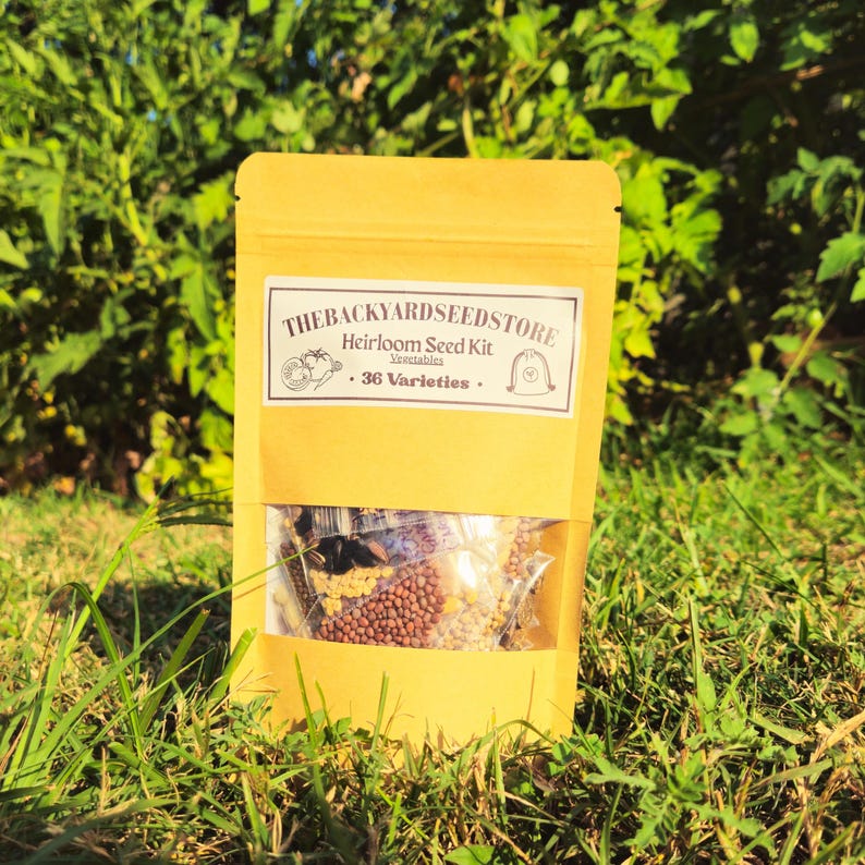 May include: A yellow paper bag with a clear window, filled with seeds. The bag has a white label that reads "THEBACKYARDSEEDSTORE Heirloom Seed Kit Vegetables . 36 Varieties ." The bag is sitting on green grass.