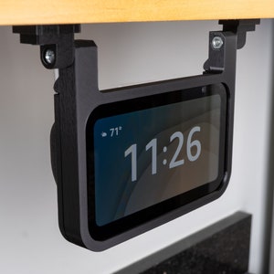 May include: A black phone holder with a phone displaying the time 11:26 and the temperature 71 degrees. The holder is mounted under a wooden shelf.
