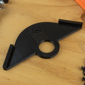 May include: Black plastic jig with a 6.35 cm diameter hole and two angled wings. The jig is designed to help with woodworking projects.