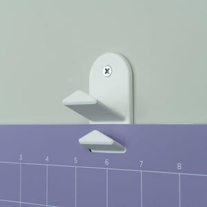 May include: White plastic wall mount with a hook for hanging a calendar or other items. The mount is attached to a wall with a screw. The calendar is a light purple color with numbers 1 through 9 printed on it.