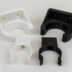 May include: Four plastic wall mounts, two white and two black, designed to hold pipes or other cylindrical objects. The mounts have a U-shaped opening and two holes for attaching to a wall.