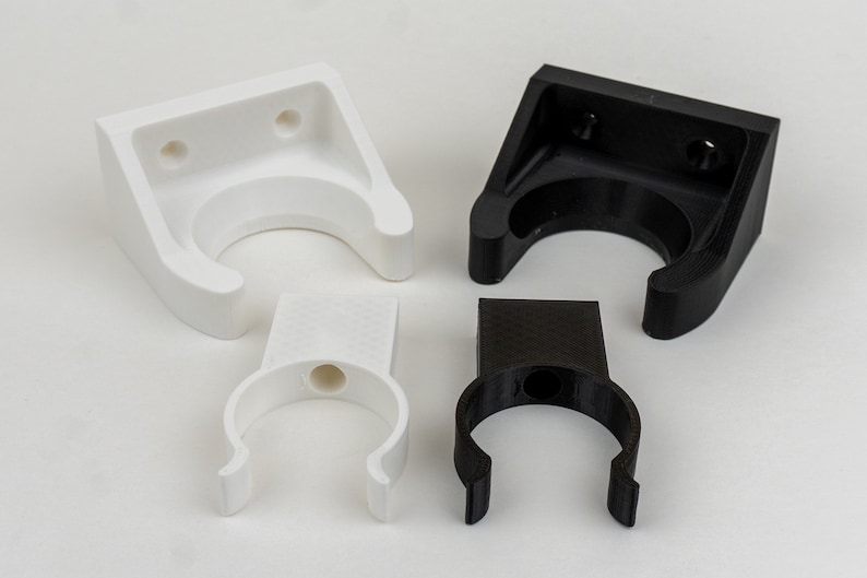 May include: Four plastic wall mounts, two white and two black, designed to hold pipes or other cylindrical objects. The mounts have a U-shaped opening and two holes for attaching to a wall.