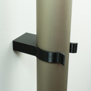 May include: A black plastic wall mount for holding a round object, such as a pipe or rod. The mount has a curved design to securely hold the object in place.