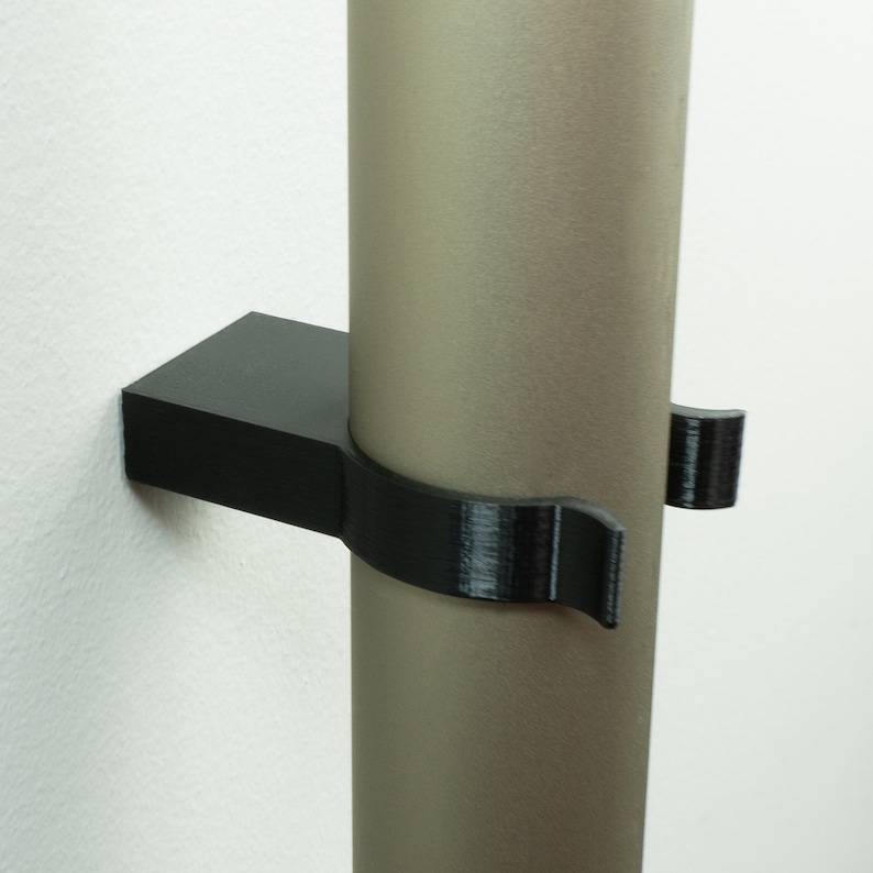 May include: A black plastic wall mount for holding a round object, such as a pipe or rod. The mount has a curved design to securely hold the object in place.
