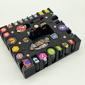 Könnte beinhalten: Schwarzer Plastik-Organizer mit Fächern zur Aufbewahrung von kleinen Spielsteinen. Der Organizer enthält eine Vielzahl von bunten Spielsteinen mit verschiedenen Designs und Symbolen.