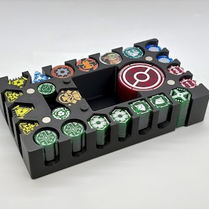 Op de afbeelding: Zwarte organizer met meerdere compartimenten voor spelonderdelen. De organizer bevat stapels groene en rode fiches, evenals ronde fiches met verschillende ontwerpen. Het ontwerp omvat een centraal gebied voor een stapel rode fiches.