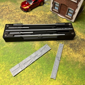 Puede incluir: Una caja rectangular negra con varios componentes rectangulares grises. Dos piezas rectangulares grises con diseños grabados están separadas. Un coche de juguete rojo y un edificio en miniatura en el fondo.