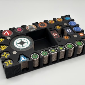 May include: Black organizer for board game components. Features various compartments for tokens and cards. Includes a central storage area and slots for stacks of hexagonal tokens. The organizer has a variety of colorful game pieces.