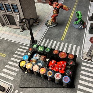 May include: A black plastic organizer with compartments for dice and tokens. The organizer has a variety of colorful tokens and dice, including red dice, green tokens, and blue tokens. The organizer is sitting on a table with a black and white checkered pattern.