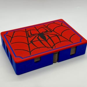 May include: A rectangular blue storage box with a red lid featuring a black spider and web graphic. The box has cutouts on the sides revealing stacked cards inside. The lid is bordered by a thin blue line, and a small spider web is embossed on the side.