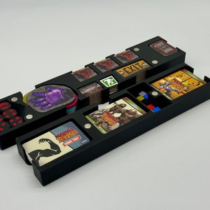 May include: Black plastic organizer with compartments for storing a board game. The organizer has a compartment for dice, a compartment for cards, and a compartment for tokens. The organizer is labeled "Marvel Zombies:  Heroic Trait".