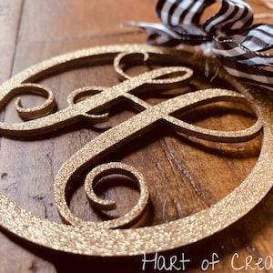 May include: Gold glitter monogram ornament with a black and white ribbon bow. The monogram is the letter "F" in a script font.