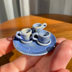 Puede incluir: Miniatura de juego de té de cerámica azul con tres tazas y platillos sobre un plato a juego. El juego tiene una apariencia rústica y hecha a mano con un esmalte moteado. El juego se sostiene en una mano, mostrando su pequeño tamaño.