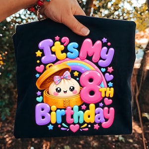 Puede incluir: Una camiseta negra con un gráfico colorido de una bola de masa sonriente en una vaporera de bambú, un arcoíris y el texto "It's My 8th Birthday" en letras de burbuja. El diseño incluye pequeñas estrellas y corazones de colores.