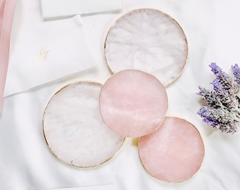 May include: A collection of rose quartz coasters with gold trim, a white pouch with a pink ribbon, a white card with gold text, and a small bundle of lavender on a white surface. The card reads "S.Theory Bringing aesthetic to life."