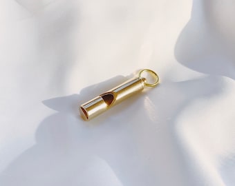 May include: A small, gold-colored whistle with a ring for attachment. The whistle is cylindrical with a narrow opening and a small hole. It is resting on a white, silky fabric.