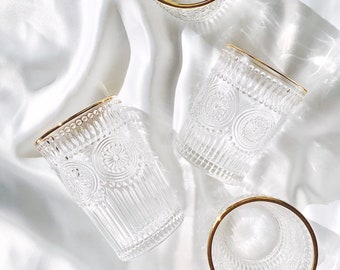 May include: Four clear drinking glasses with gold rims are arranged on a white, silky surface. The glasses have a detailed, embossed pattern. The light creates shadows and highlights on the glasses and the fabric.
