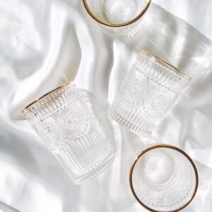 May include: Four clear drinking glasses with gold rims are arranged on a white, silky surface. The glasses have a detailed, embossed pattern. The light creates shadows and highlights on the glasses and the fabric.