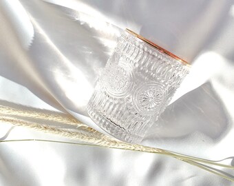 May include: A clear, patterned glass with a gold rim sits on a white, silky surface. The glass features an intricate design of repeating circular patterns. Dried wheat stalks are placed near the glass, adding a natural element to the composition.