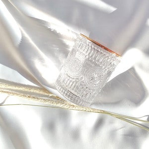 May include: A clear, patterned glass with a gold rim sits on a white, silky surface. The glass features an intricate design of repeating circular patterns. Dried wheat stalks are placed near the glass, adding a natural element to the composition.