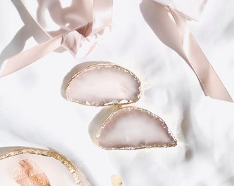 May include: Two agate coasters with gold edges and a pale pink hue, alongside a matching ribbon and a decorative plate with dried petals. The items are arranged on a white, silky surface, creating a soft, elegant aesthetic.