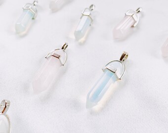 May include: A collection of crystal pendants in various colors, including pink, white, and blue. Each pendant has a hexagonal shape and is capped with a silver-colored metal bail for hanging. The pendants are arranged on a white fabric surface.