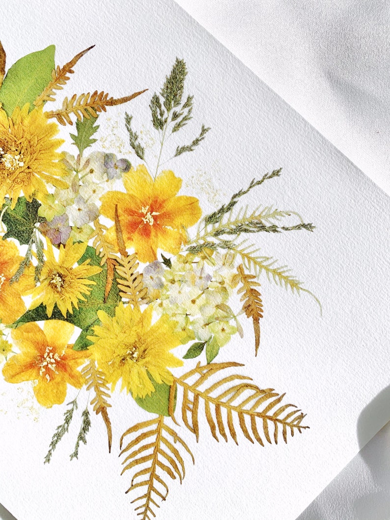 Pu&ograve; includere: Primo piano di una composizione floreale con fiori gialli e arancioni, foglie verdi e felci secche. I fiori sono disposti su una superficie bianca strutturata, creando una composizione naturale e artistica.