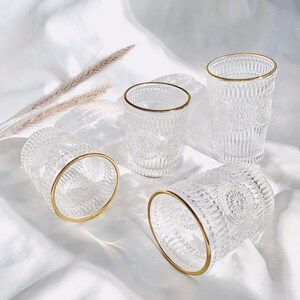 May include: Four clear drinking glasses with gold rims. The glasses have a detailed, embossed pattern. The glasses are arranged on a white, silky surface. The glasses are empty.