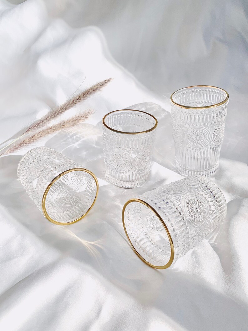 May include: Four clear drinking glasses with gold rims. The glasses have a detailed, embossed pattern. The glasses are arranged on a white, silky surface. The glasses are empty.