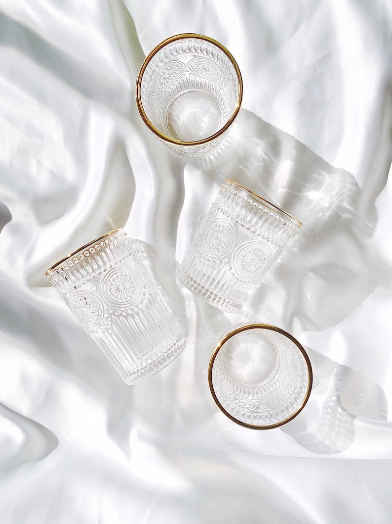 May include: Four clear drinking glasses with gold rims are arranged on a white, silky surface. The glasses have a detailed, embossed pattern. The light creates shadows and highlights on the glasses and the fabric.