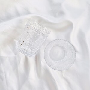 May include: Two clear glass drinking glasses with embossed floral patterns. One glass is upright, and the other is on its side. The glasses are on a white, textured surface. The glasses are likely for water or other beverages.